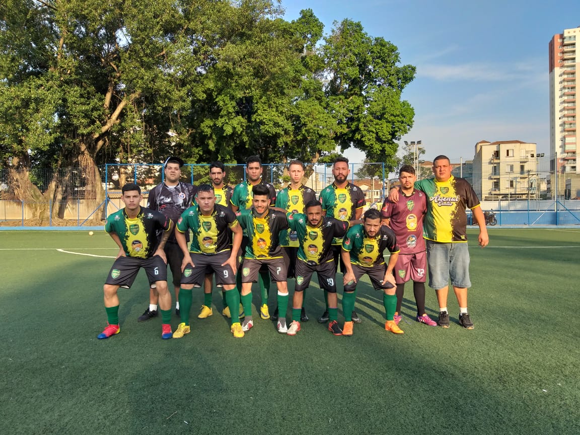 Fotografia colorida mostra time Arsenal do Centro em pose para foto em campo de futebol. Uniforme é uma camisa verde limão com golas verde escuro e bermudas pretas. 